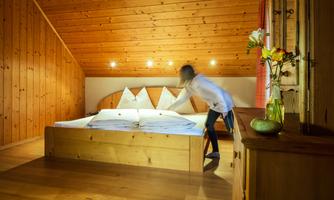 A cozy bedroom with wooden paneling and a large bed. One person is arranging the bedding and there are fresh flowers on a table.