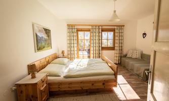 A bright bedroom with a wooden bed and a cozy sofa bed. Large windows let in plenty of light and offer a view of the outdoors.