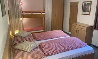 A simple bedroom with two single beds and a loft bed in the background. There is a wardrobe and a cozy atmosphere.