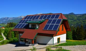 A modern house with solar panels on the roof.  
Surrounded by green meadows and mountains in the background.