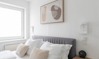 A modern bedroom with a gray bed and white pillows. An abstract artwork hangs on the wall, and there is a wooden nightstand with a clock.