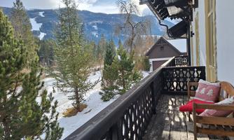 A balcony overlooking snow-covered mountains and coniferous trees. In the foreground, there is a cozy chair with a red cushion.