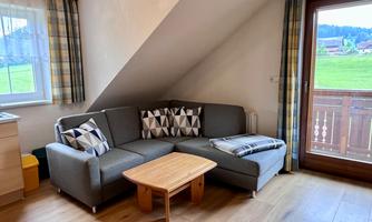 A cozy living room with a gray sofa and a small wooden table. Large windows offer a view of the greenery.
