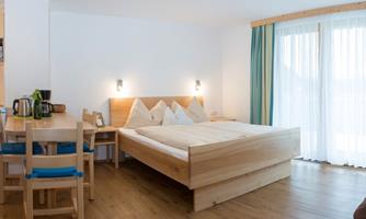 A bright room with a large double bed and wooden furniture. There are chairs at the table and a view of the balcony through the windows.