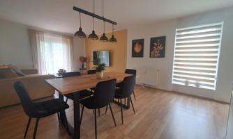 A modern dining room with a long table and black chairs. In the background, there is a comfortable sofa and large windows with blinds.