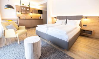 A modern room with two beds, a cozy armchair, and a small kitchenette. The wooden elements give the space a warm atmosphere.