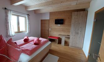A cozy bedroom with a double bed, red pillows, and wooden ceilings. There is a television and a small table in a rustic style.