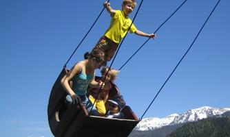 A group of children is playing on a large swing with a view of the mountains. The sky is clear and blue.