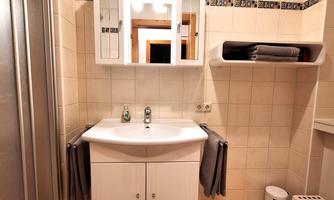 A modern bathroom with a white sink and a mirror. The walls are tiled and there is a shower cabin as well as a towel holder.
