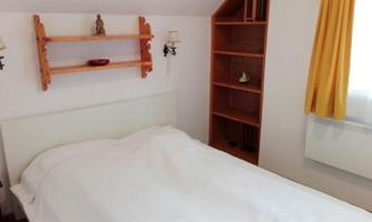A simple bedroom with a bed and a bedside table. On the wall there are shelves and a large window with yellow curtains.