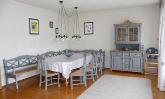 A bright dining room with a long table and six chairs. Pictures hang on the wall and there is a wooden cabinet in the room.