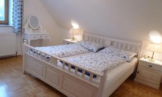 A bright bedroom with a large white bed and two bedside tables. The room has wooden flooring and a window that lets in daylight.