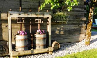 A rustic wooden building with old beverage presses and colorful flowers in barrels. The garden is green and well-maintained, with gravelly ground.