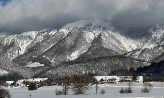 A winter landscape with snow-covered mountains and a clear sky. In the foreground, there are some houses and trees visible.