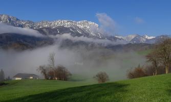 A picturesque landscape with green meadows and mountains in the background. Gentle fog drifts through the valley under a clear blue sky.