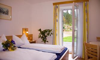 A cozy bedroom with two beds and a window opening to the garden. A plant arrangement and bright light create an inviting atmosphere.