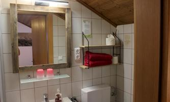 A modern bathroom with bright tiles and wooden details.  
On the sink, there are two red towels, and decorative elements are arranged on a shelf.