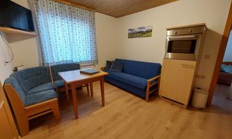 A cozy lounge with a blue couch, a table, and a cabinet. Bright walls and a window provide friendly light.