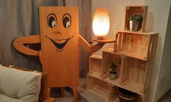 A cheerful figure in the shape of a box stands next to a shelf made of wooden crates. In the background, a lamp glows and there are some plants on the shelf.