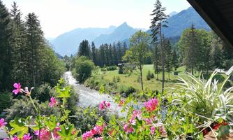 A picturesque view of a river, surrounded by green meadows and tall trees. In the foreground, colorful plants are blooming.