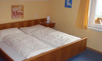 A simple bedroom with a double bed and yellow walls. The room has a table, windows with curtains, and atmospheric wall pictures.