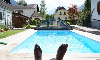 A swimming pool with clear water and a glass roof. In the foreground, feet can be seen relaxing on a deck chair.