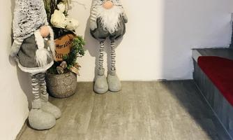 Two decorative figures in the form of gnomes stand in a cozy entrance area. In front of them lies a doormat with the inscription "How nice that you are here."