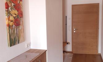 A bright hallway with wooden flooring and a cozy dresser. A colorful floral painting hangs on the wall.
