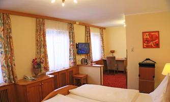 A cozy hotel room with a double bed, a desk, and a television. The walls are light-colored, and the windows have colorful curtains.