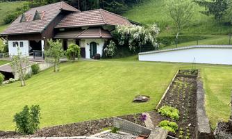A beautiful house with a well-maintained garden and a green lawn. In the background, there are trees and hills visible.