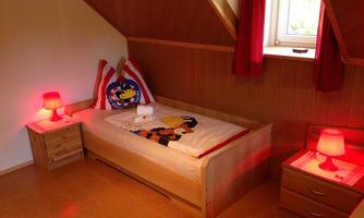 A cozy bedroom with a single bed and colorful lamps. The walls are made of wood and a window provides daylight.