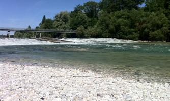 A clear river with a rocky shore and gentle waves. In the background, a bridge and lush trees can be seen.