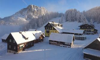A picturesque alpine landscape with snow-covered huts and mountains in the background. The clear sky and the snow create a tranquil winter atmosphere.