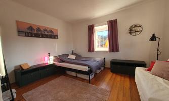 A bright bedroom with a bed and a cozy sofa. The walls are painted light and a rug is on the wooden floor.