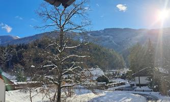 A snowy winter landscape with a large tree and mountains in the background. The sun shines brightly in the sky, creating a peaceful atmosphere.