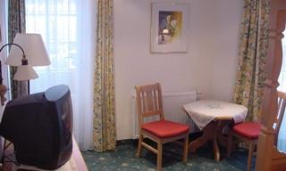 A cozy living area with a table and two chairs. In the background, there is a television and there are windows with curtains.