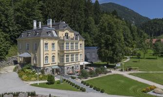 A historic, elegant building surrounded by green grass and trees. Mountains are visible in the background and the sky is clear.