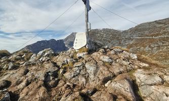 A summit cross on a rocky mountain peak, surrounded by an impressive mountain landscape. The sky is clear with some clouds.
