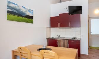A modern kitchen with red cabinets and a wooden table. On the wall hangs a picture of a green landscape under a cloudy sky.