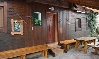 A cozy wooden cabin with a brown facade and a rustic entrance. In front of the cabin are long benches and a table, surrounded by green plants.