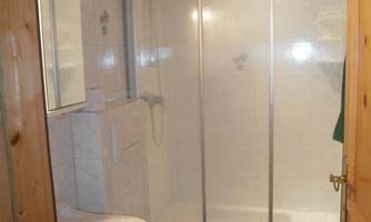 A small bathroom with a shower, a sink, and a toilet. The walls are tiled in light colors, and there is a wooden ceiling.