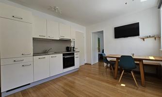 A modern kitchen with white cabinets, a stove, and a refrigerator. In the background, there is a wooden table with chairs and a television on the wall.