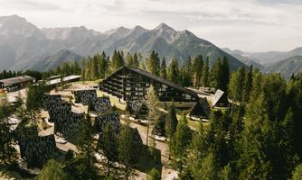 A modern hotel building surrounded by green trees and mountains. The architecture harmoniously blends with nature.