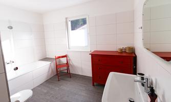 A modern bathroom with white tiles and a red chair. There is a window, a bathtub, and a sink.