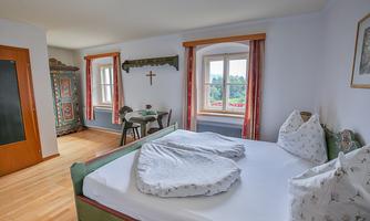 A cozy bedroom with a large bed, a wooden table, and chairs. The windows offer a view of greenery and the walls are painted in light colors.