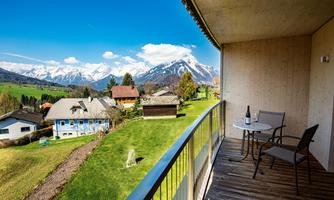 A beautiful view from a balcony over green meadows and snow-covered mountains. In the foreground, there is a table and a chair, perfect for relaxing.