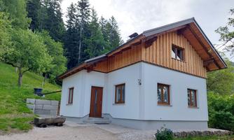 A modern house with wooden cladding in a green environment. It is located on a slope, surrounded by trees and meadows.