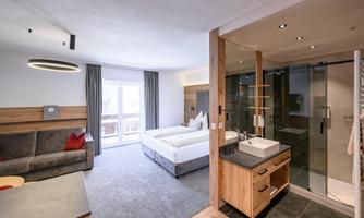 A modern room with a double bed and a cozy seating area. On the right side, there is a bathroom with a glass shower and wooden details.
