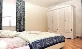 A cozy bedroom with a double bed and a white wardrobe. The windows are adorned with curtains and the floor is made of wood.