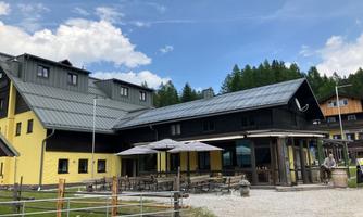 A modern building with a yellow and black painted facade. Outside, there are tables with sun umbrellas in a green environment.